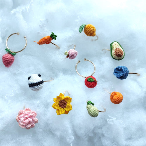 Bague en crochet fait main représentant des fruits et des fleurs colorés, dont une fraise, une carotte, un avocat, un tournesol et plus encore, sur un fond de nuages blancs