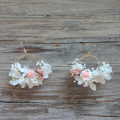 Boucles d'oreilles florales Clara avec des fleurs éternelles blanches et roses, posées sur un fond en bois naturel.
