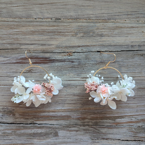 Boucles d'oreilles florales Clara avec des fleurs éternelles blanches et roses, posées sur un fond en bois naturel.
