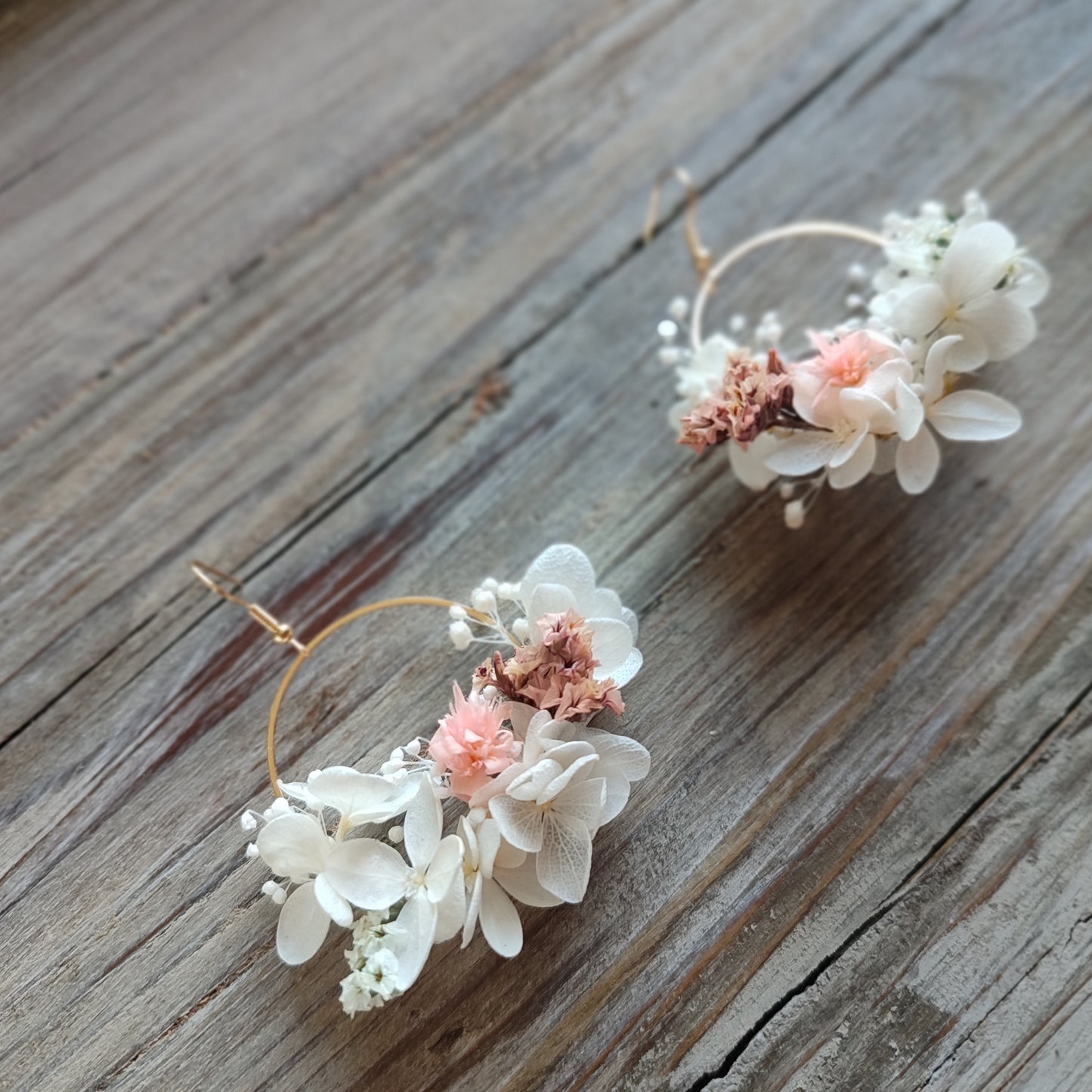 Boucles d'oreilles florales Clara avec des fleurs éternelles blanches et roses, posées sur un fond en bois naturel.