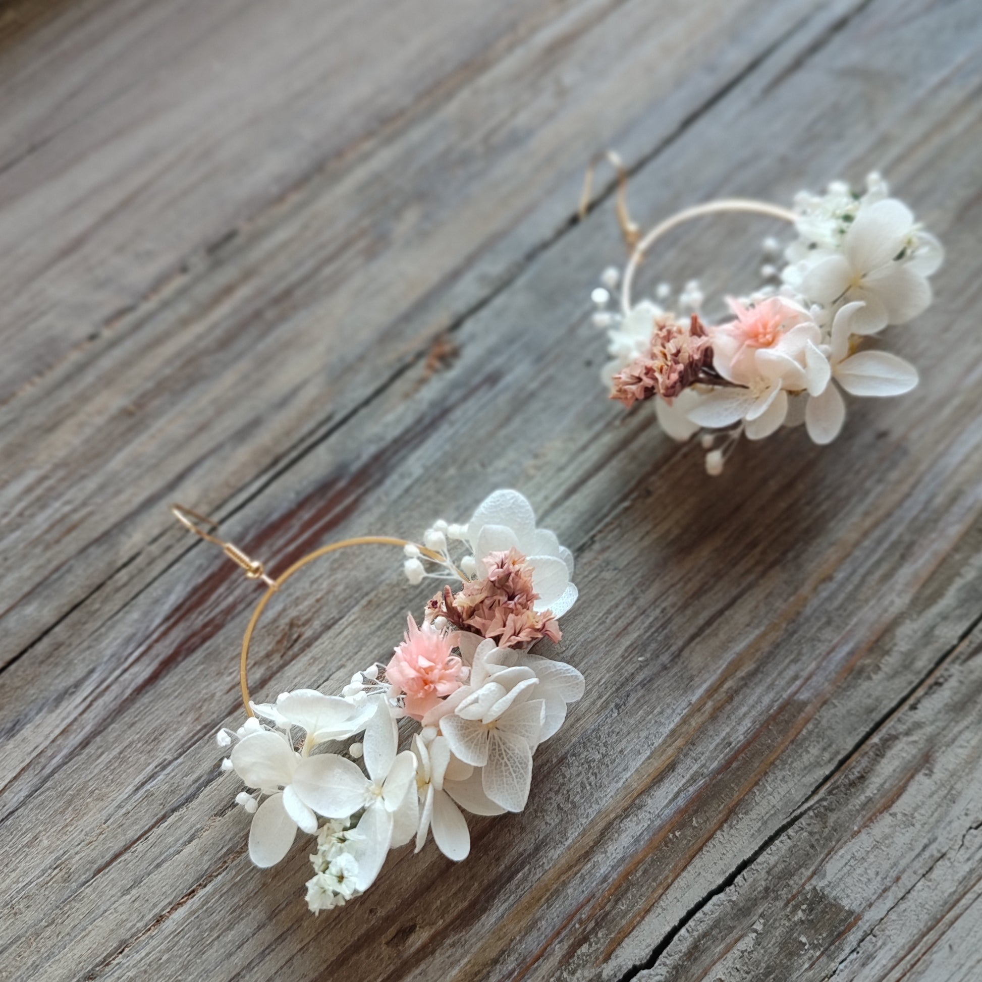 Boucles d'oreilles florales Clara avec des fleurs éternelles blanches et roses, posées sur un fond en bois naturel.