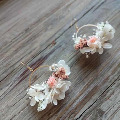 Boucles d'oreilles florales Clara avec des fleurs éternelles blanches et roses, posées sur un fond en bois naturel.
