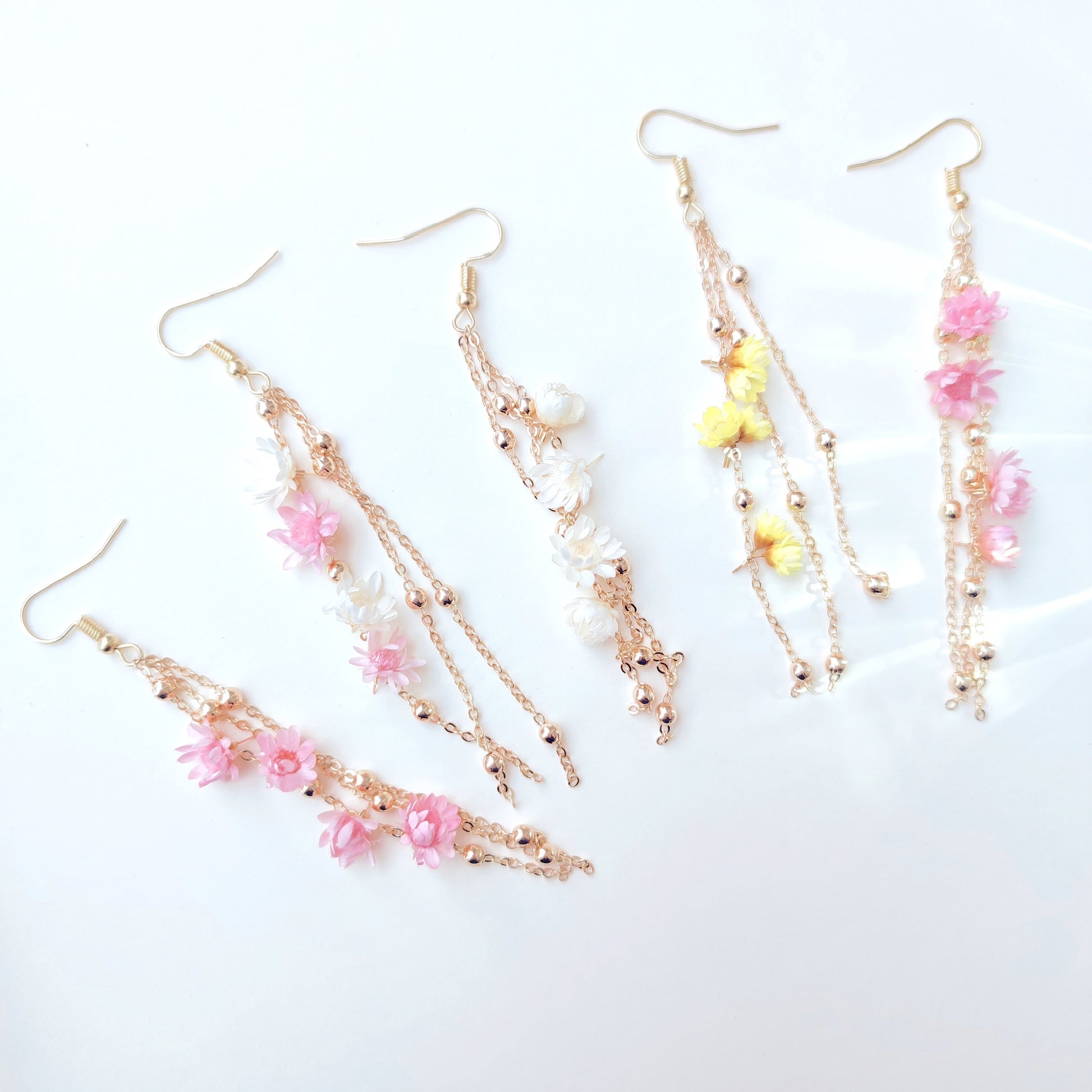 Boucles d’oreilles Élise en fleurs séchées, ornées de chaînes dorées et de perles délicates, disponibles en blanc, rose, et jaune.