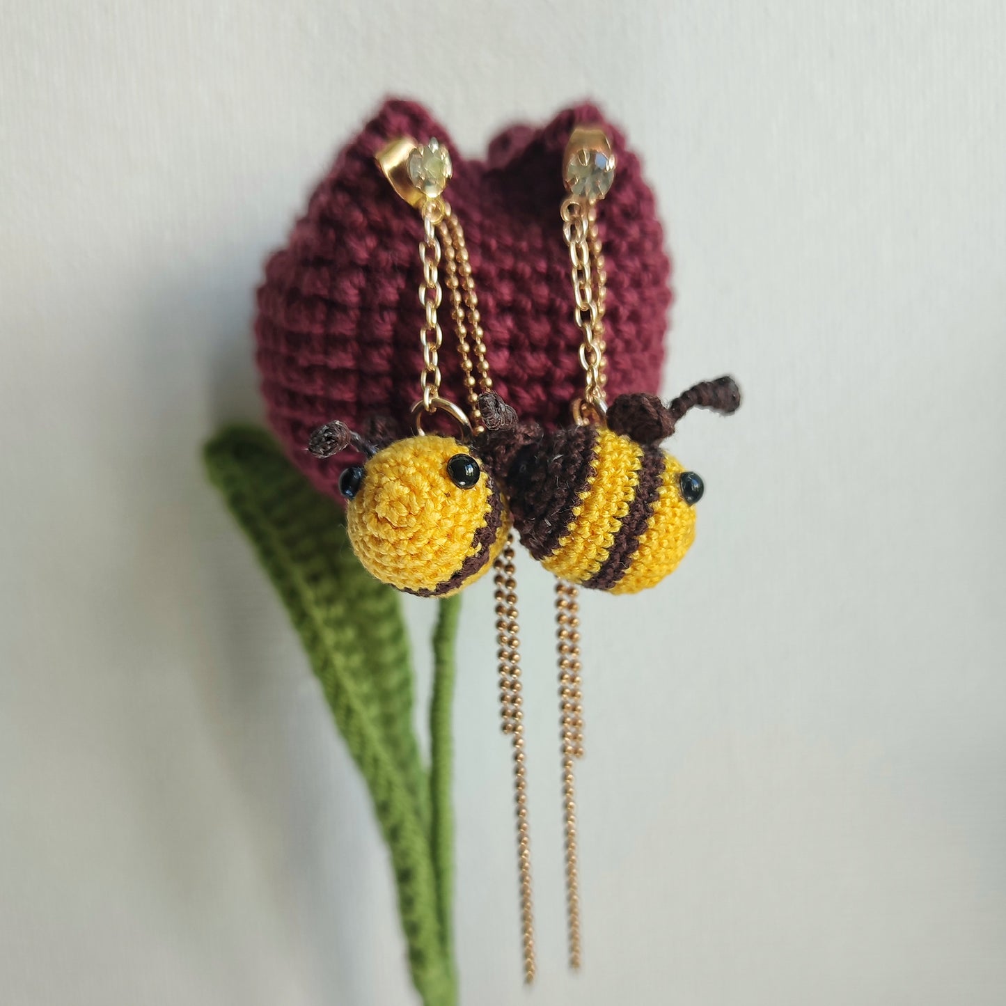 Deux boucles d'oreilles abeille en crochet, une suspendue à une chaîne dorée et l'autre à une tulipe crochetée.