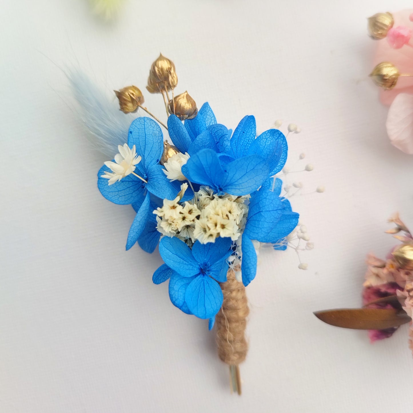 Boutonnière florale bleue avec des fleurs préservées et des accents dorés.
