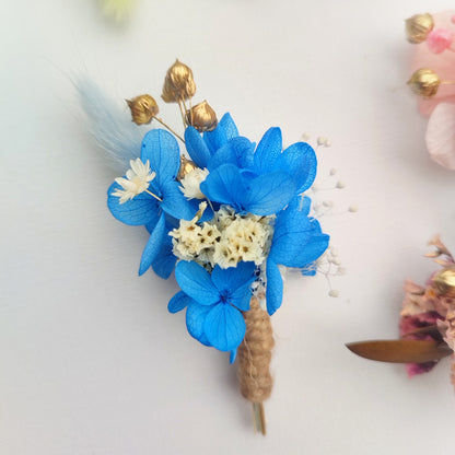 Boutonnière florale bleue avec des fleurs préservées et des accents dorés.