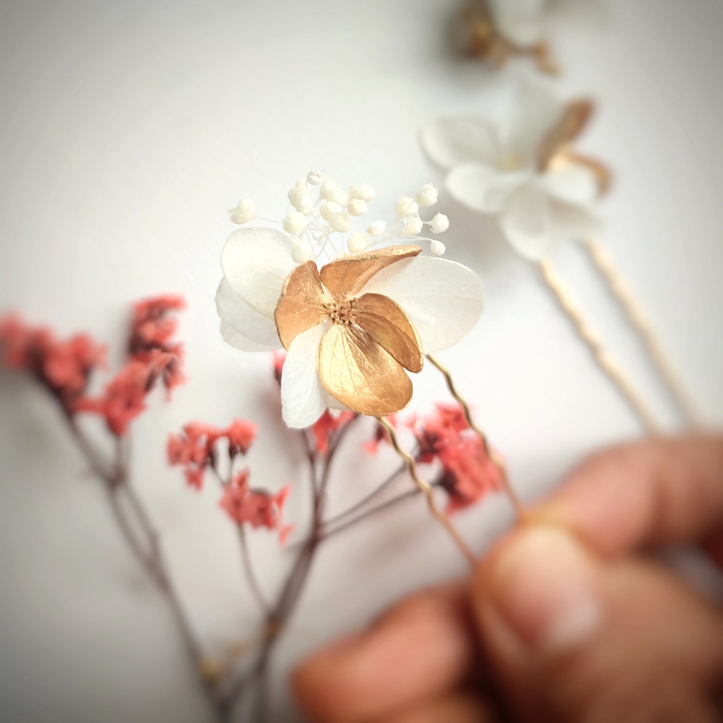 Détail des épingles à cheveux "Éclat Floréal Alcina" montrant les fleurs stabilisées blanches et dorées et leur finition artisanale.
