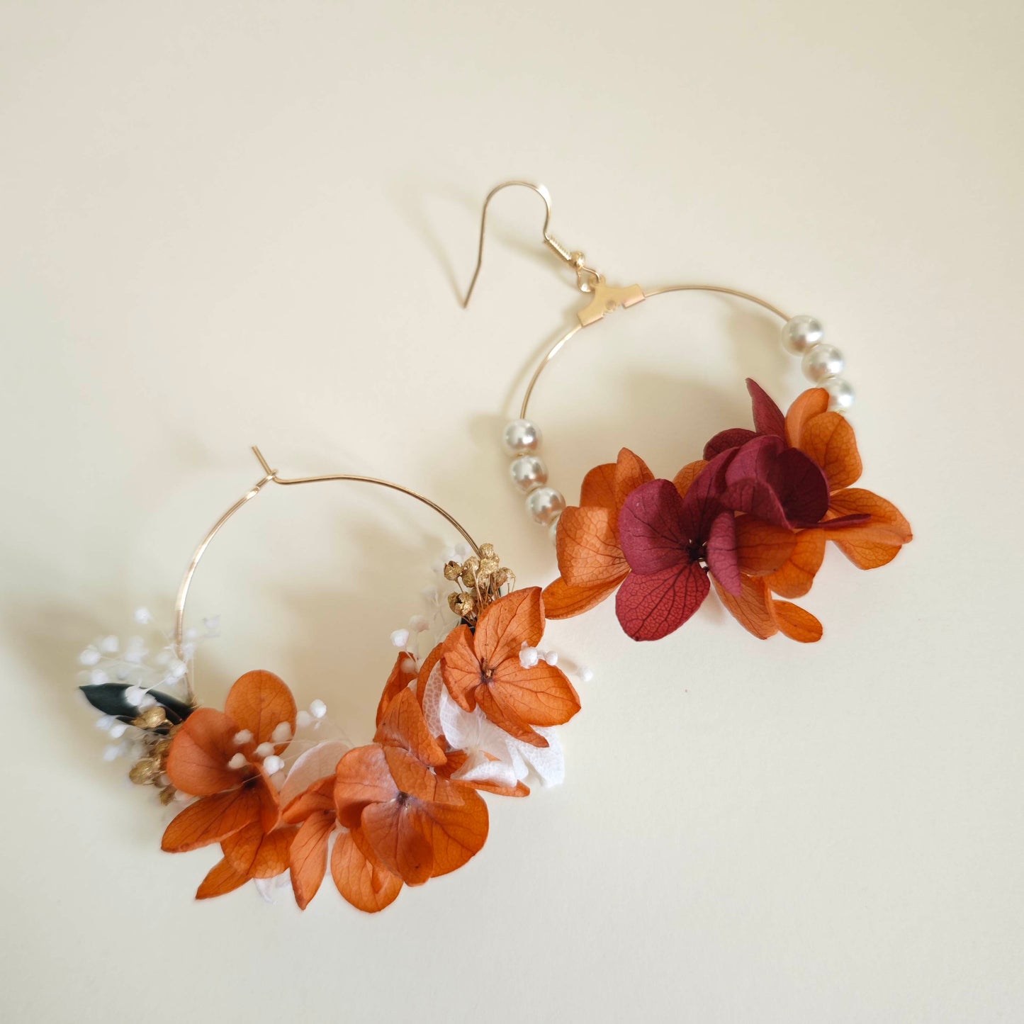 Paire de boucles d’oreilles créoles faites main, ornées de fleurs séchées orange, blanches et bordeaux, avec perles blanches sur un fond beige.