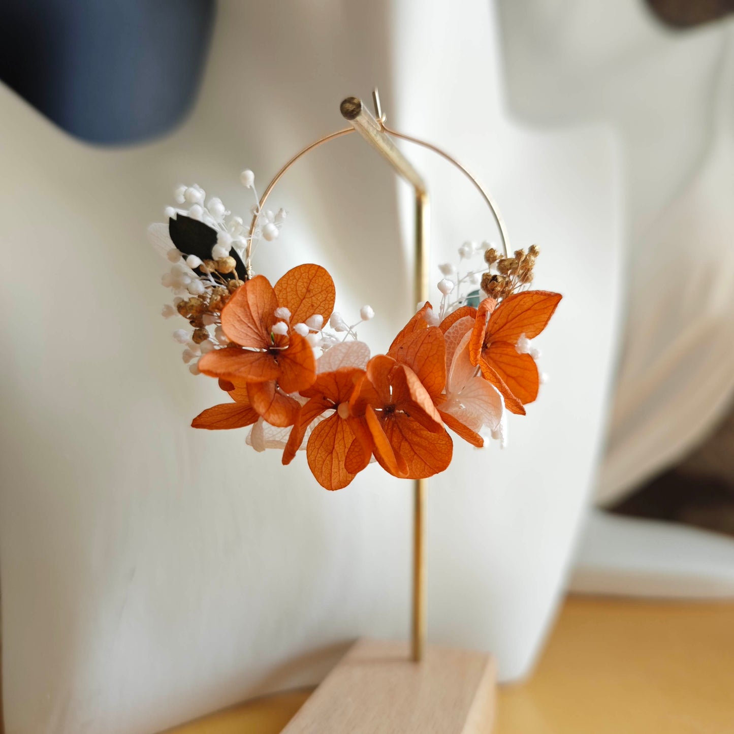 Boucle d’oreille en fleurs séchées orange et blanches portée sur un buste blanc, devant un mur en pierre naturelle.