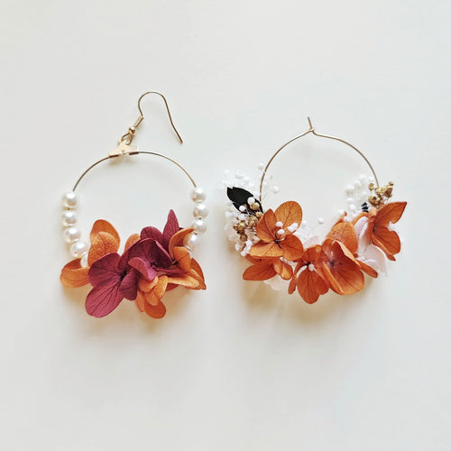 Paire de boucles d’oreilles créoles faites main, ornées de fleurs séchées orange, blanches et bordeaux, avec perles blanches sur un fond beige.