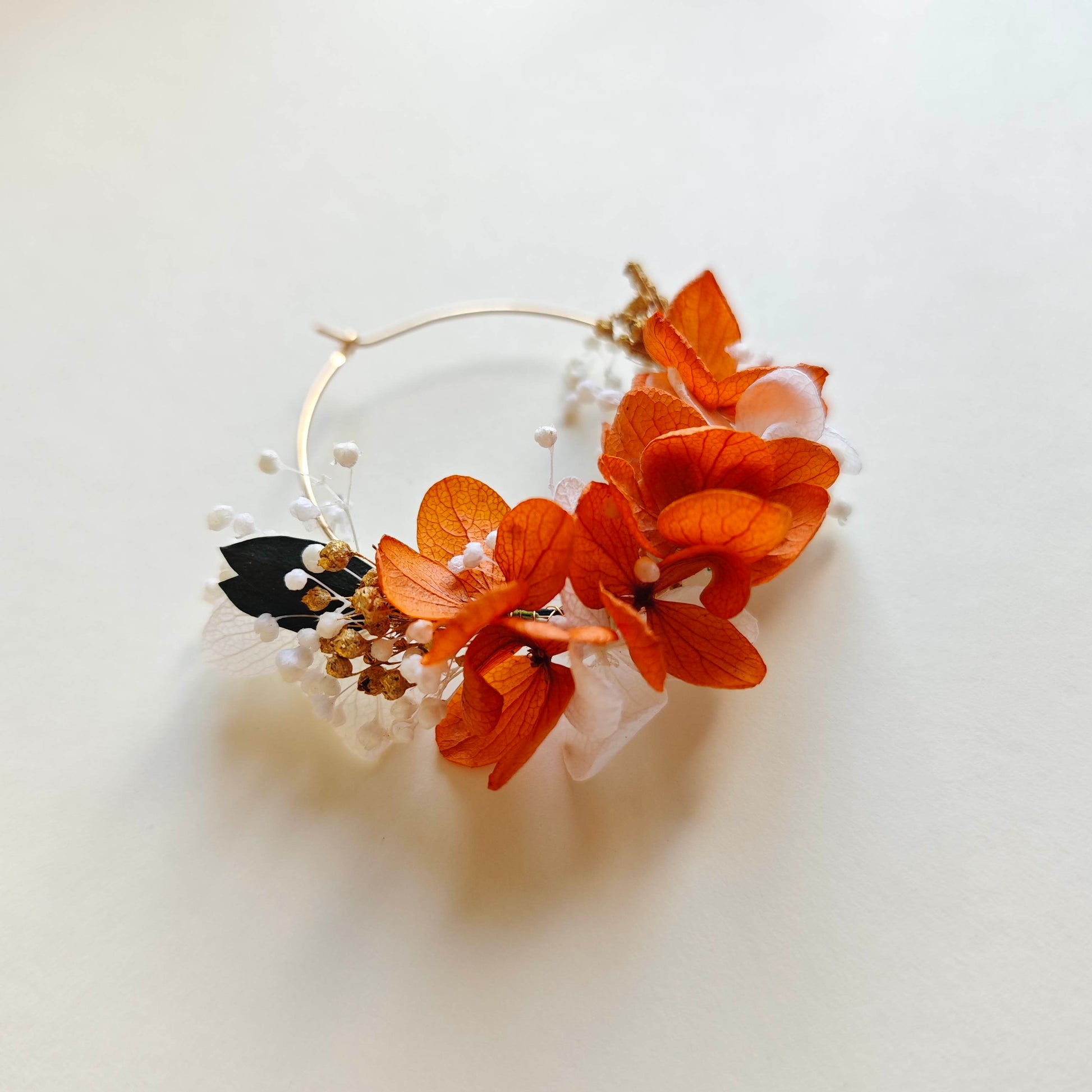 Boucle d’oreille florale posée à plat sur fond clair, composée de fleurs séchées orange, blanches et noires avec perles de gypsophile