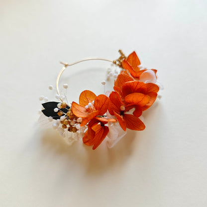 Boucle d’oreille florale posée à plat sur fond clair, composée de fleurs séchées orange, blanches et noires avec perles de gypsophile