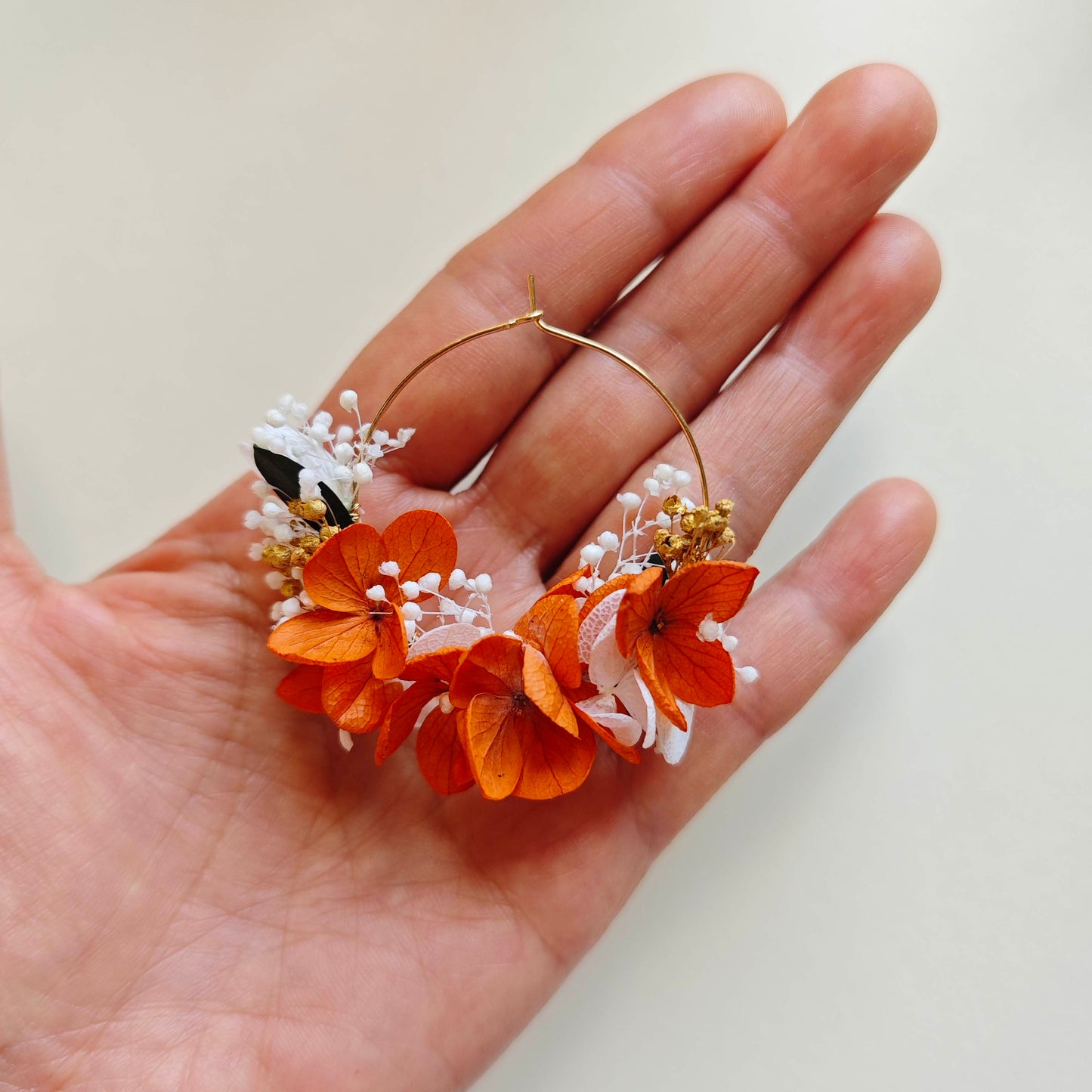 Boucle d’oreille créole faite main avec fleurs séchées orange et blanches, perles de gypsophile, sur fond de main humaine.