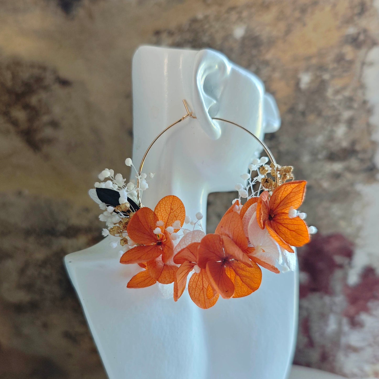 Boucle d’oreille en fleurs séchées orange et blanches portée sur un buste blanc, devant un mur en pierre naturelle.