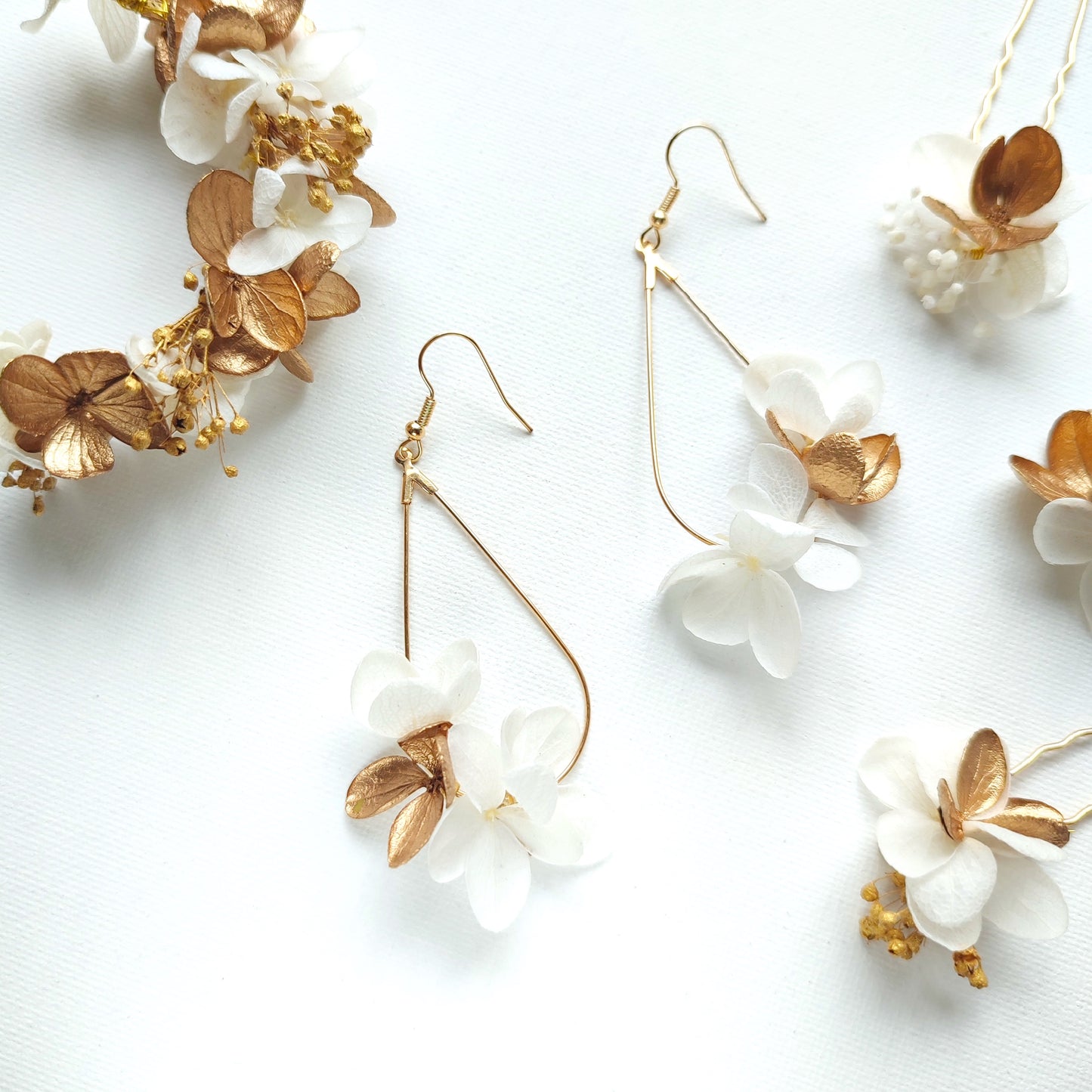 Boucles d'oreilles en forme de goutte d'eau avec des fleurs stabilisées blanches et dorées, accompagnées d'une couronne florale assortie sur un fond blanc.
