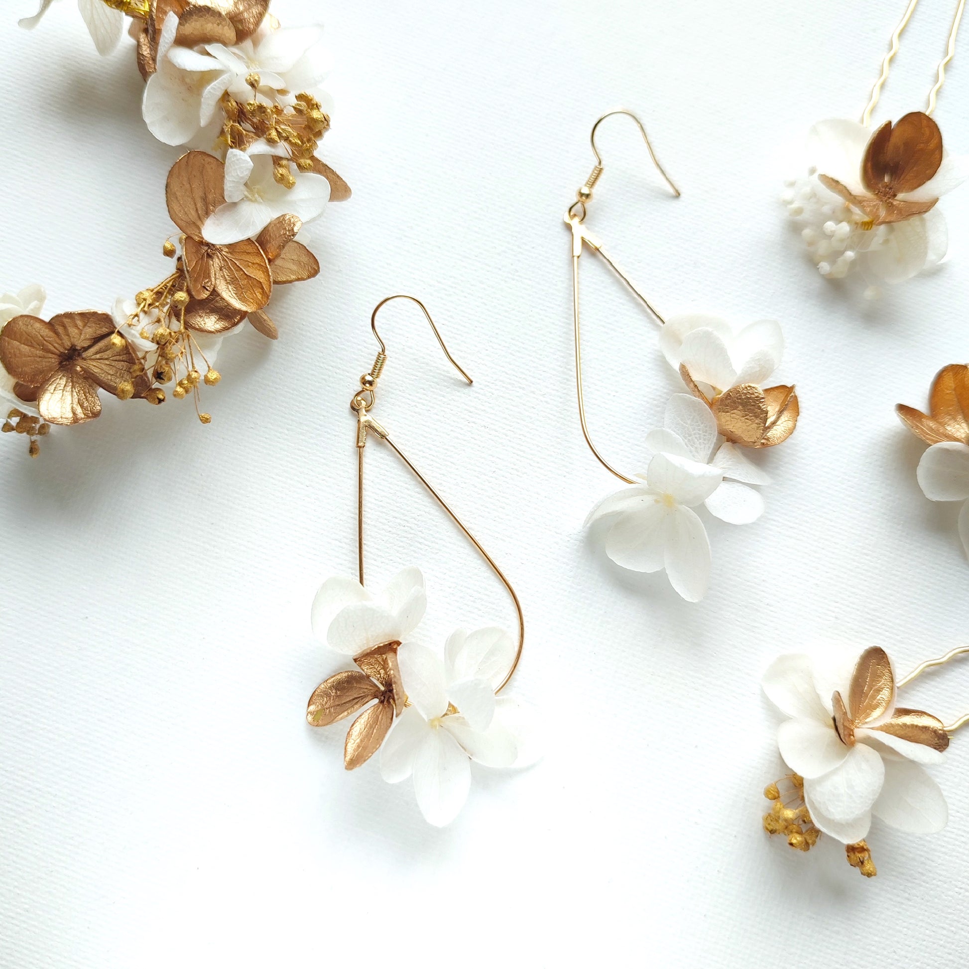 Boucles d'oreilles en forme de goutte d'eau avec des fleurs stabilisées blanches et dorées, accompagnées d'une couronne florale assortie sur un fond blanc.