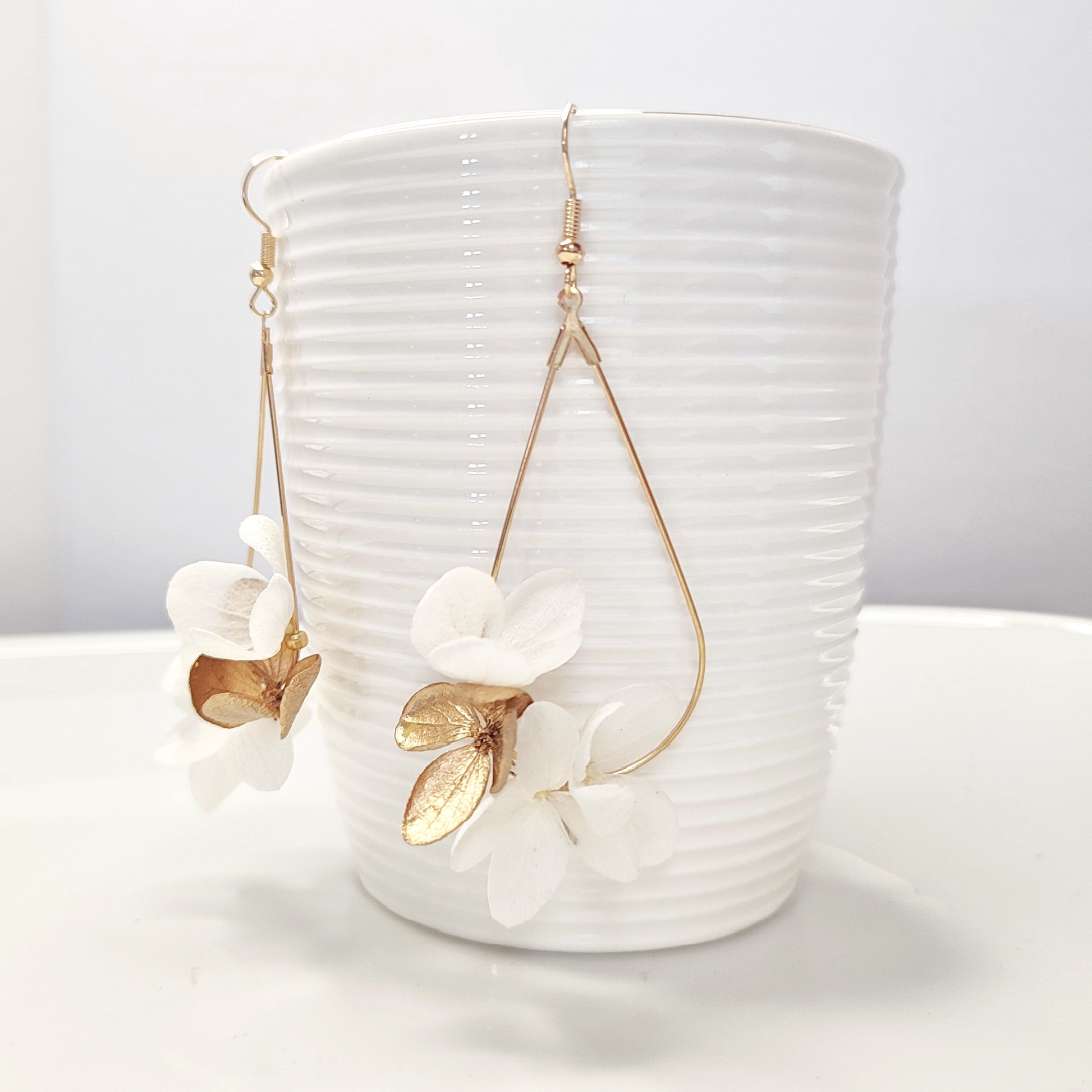 Boucles d'oreilles en fleurs stabilisées blanches et dorées, posées contre une tasse blanche côtelée, mettant en valeur leur élégance minimaliste.