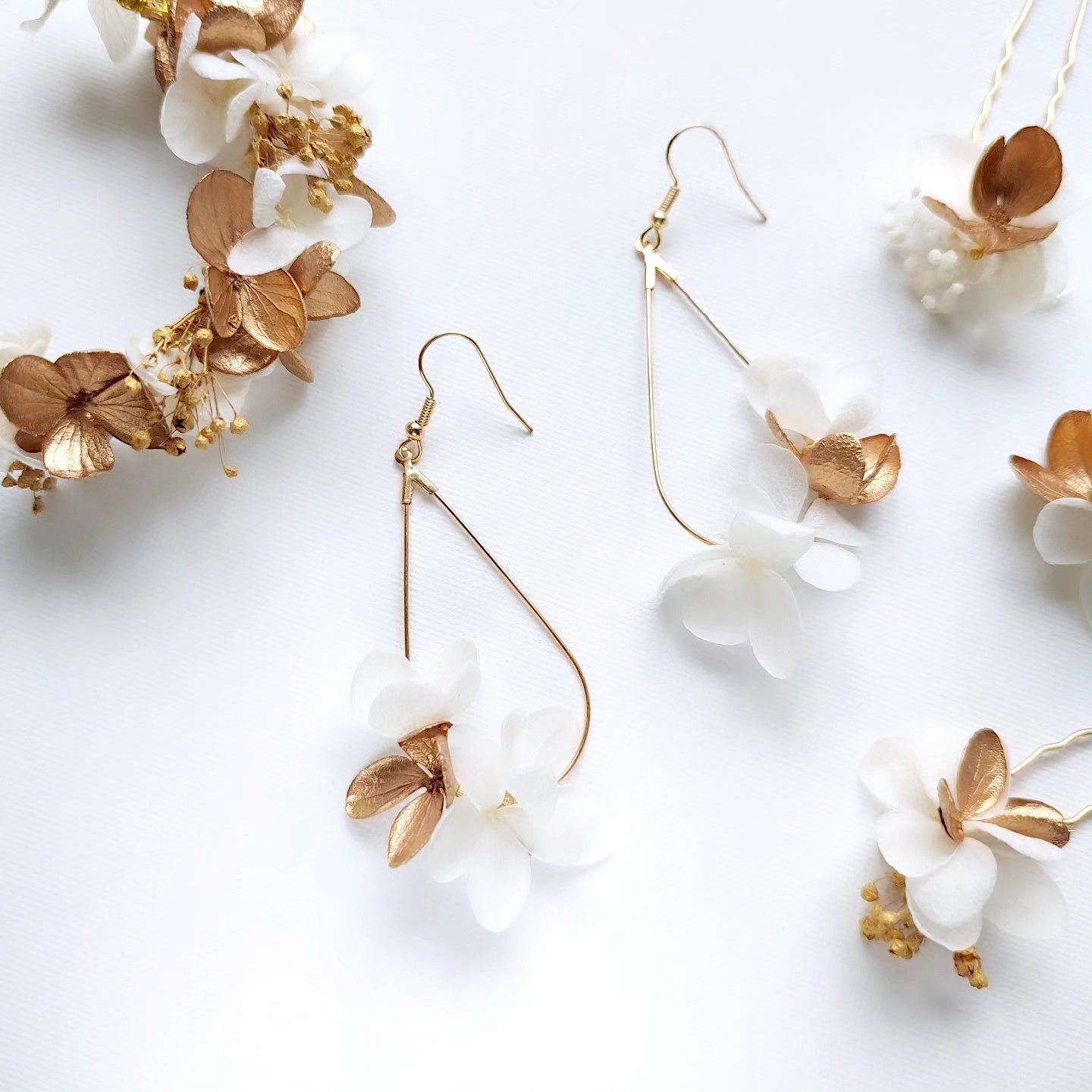 Gros plan de boucles d'oreilles en fleurs stabilisées blanches et dorées, présentées avec une couronne florale assortie et d'autres accessoires sur un fond blanc.
