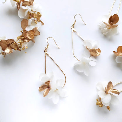 Gros plan de boucles d'oreilles en fleurs stabilisées blanches et dorées, présentées avec une couronne florale assortie et d'autres accessoires sur un fond blanc.