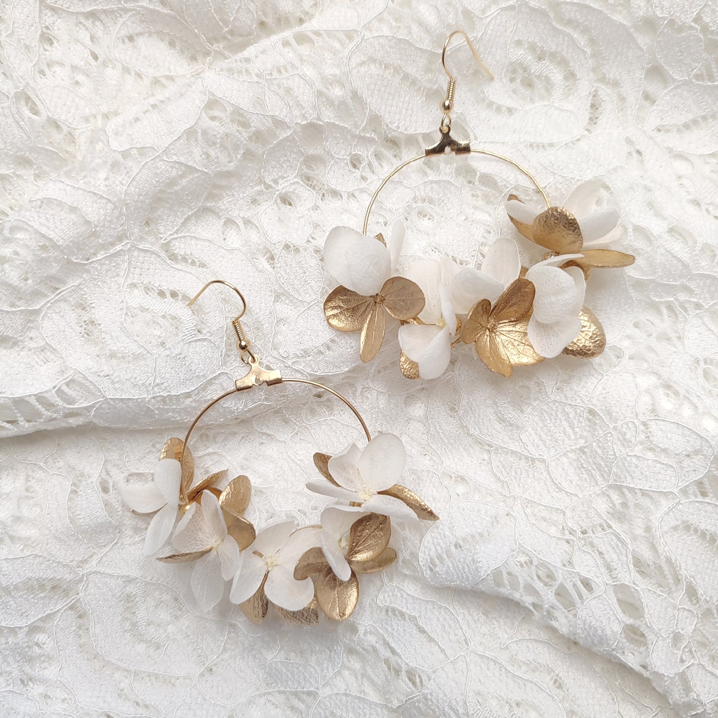 Boucles d'oreilles en créoles ornées de fleurs stabilisées blanches et dorées, posées sur un tissu blanc en dentelle, capturant l'élégance et la délicatesse des matériaux naturels.
