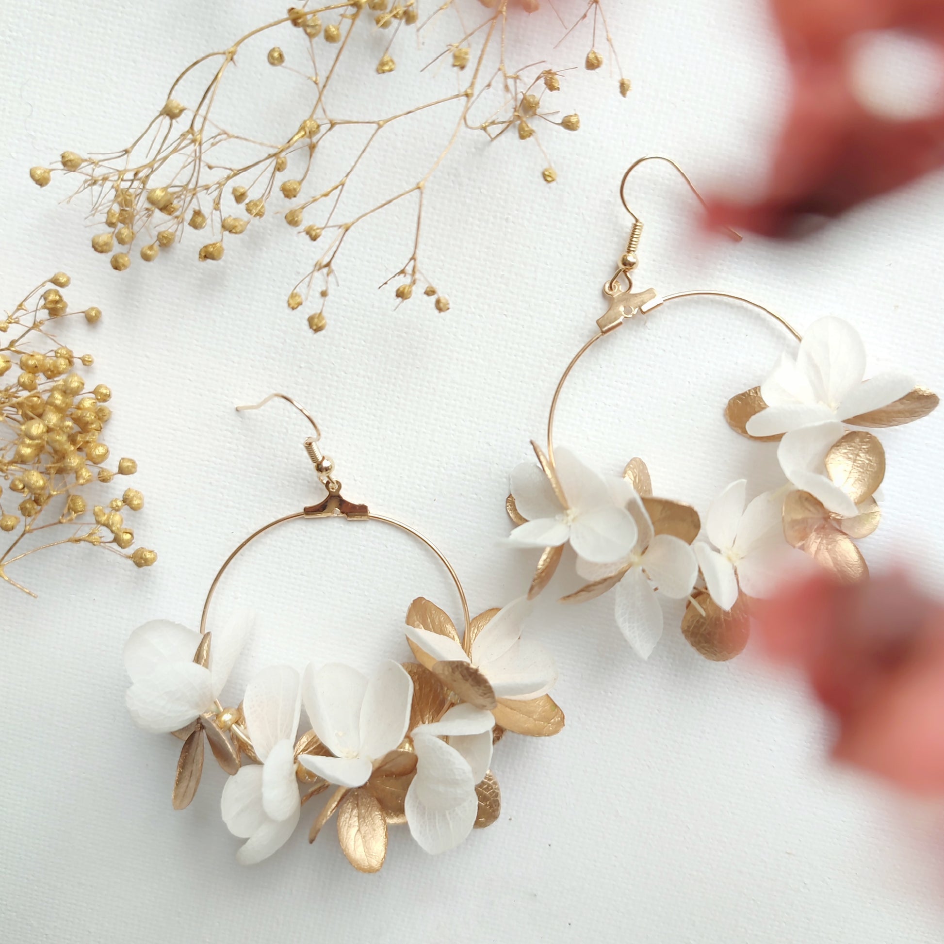 Boucles d'oreilles florales en créoles, avec des pétales stabilisés blancs et dorés, présentées sur un fond blanc avec des touches de fleurs séchées.
