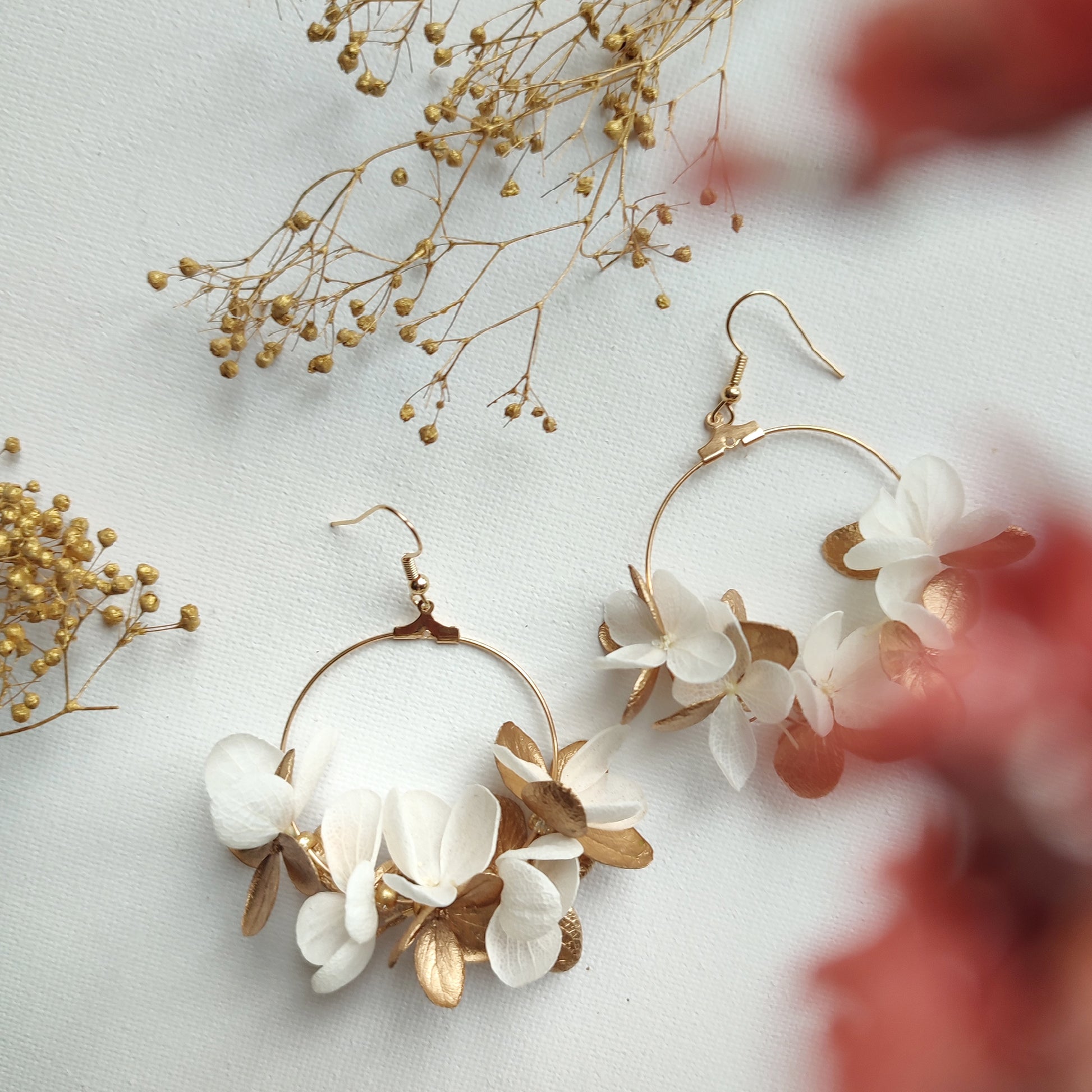 Boucles d'oreilles florales en créoles, avec des pétales stabilisés blancs et dorés, présentées sur un fond blanc avec des touches de fleurs séchées.