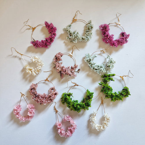 ne collection de boucles d'oreilles florales faites à la main, disposées sur une surface blanche. Les boucles d'oreilles sont disponibles en différentes couleurs, y compris  rose,blanceh , crème, rose dragée,pourpe, verte et jaune